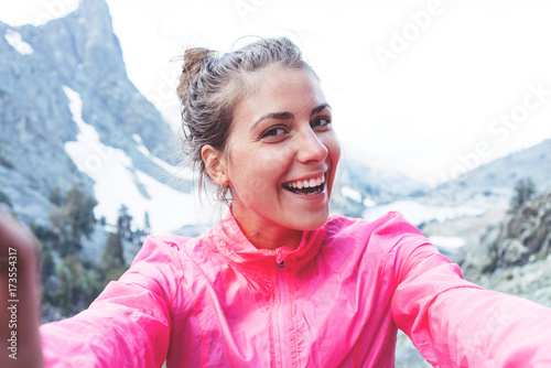 Smiling young woman taking self portrait photo high in mountains. Risky rock climbing in peaceful wilderness area. Enjoying amazing snowy lake view