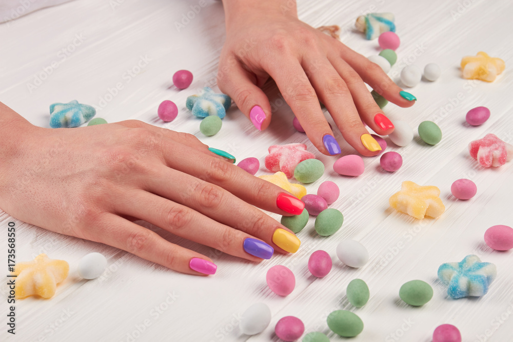 Girls manicured hands and sweets. Girls hands polished with different colors and colorful candies on white wooden background. Girls manicured hands and sweets. Girls hands polished with different colors and colorful candies on white wooden background.