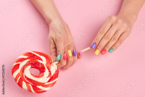 Bright lollipop in female hands. Female hands with multicolor summer nails holding tasty big lollypop on pink background, top view.
