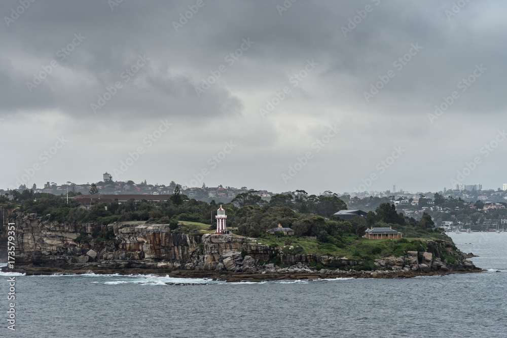 Sydney, Australia - March 21, 2017: South Head cliffs and park with short Hornby lighthouse, backed by green vegetation under foggy dark cloudscape. Part of city skyline in distance.