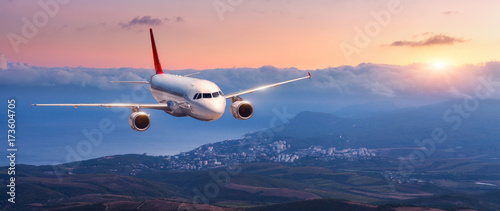 Fototapeta Samolot pasażerski. Krajobraz z białym samolotem lata w pomarańczowym niebie z chmurami nad górami, morze przy kolorowym zmierzchem. Ląduje samolot pasażerski. Samolot komercyjny. Prywatny odrzutowiec. Podróżować
