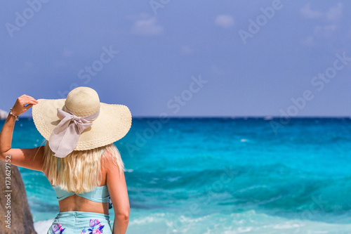 A girl in a blue summer dress with a hat on his head, standing on white sand and looking at the ocean