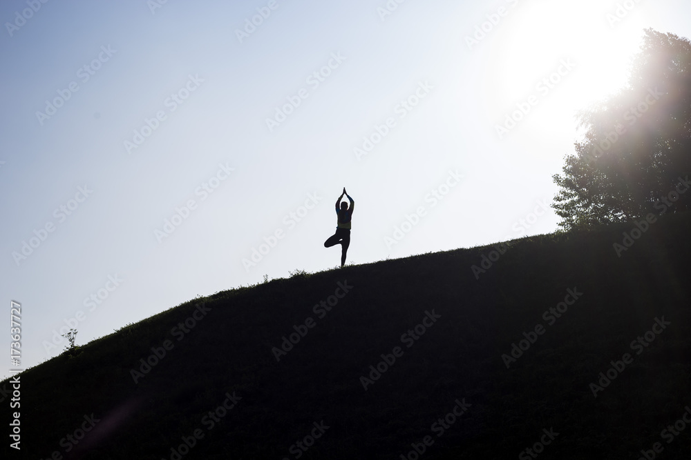 silhouette of a girl who engages in yoga on a hill in the background of a blue sky