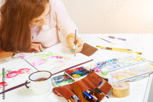 Dark-haired artist with bright draws in an album for drawing a thin wooden brush with pink watercolors, on the table lies a leather case with brushes, a mug of water, a palette of watercolors