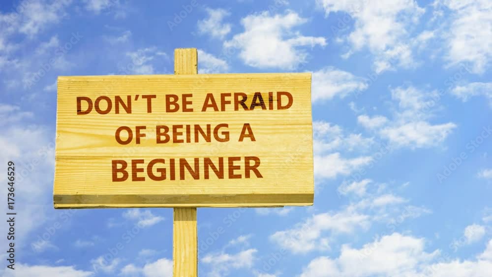 Don't be afraid of being a beginner. Words on a wooden sign against time lapse clouds in the blue sky.