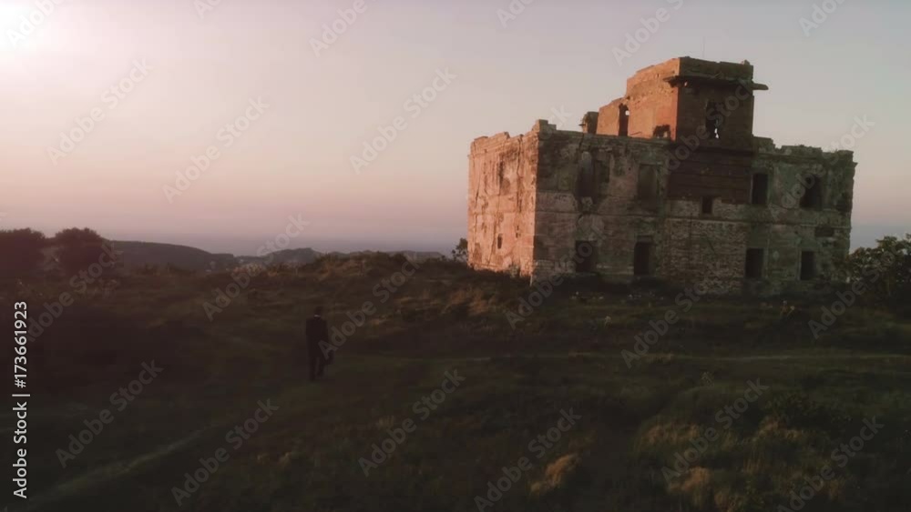 Business man in elegant suit walks toward an abandoned building in ruins at sunset on top of a mountain with the sea on the background Business man in elegant suit walks toward an abandoned building in ruins at sunset on top of a mountain with the sea on the background