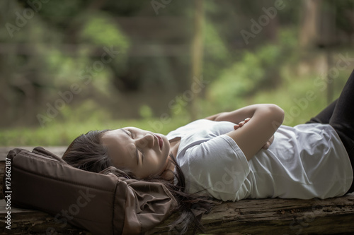 Young asian woman lay down on backpack and sleeping on wood bench in the park