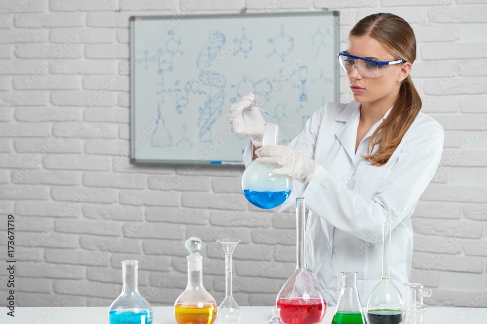 The scientist works with the chemical liquids in the laboratory. She wears protective chemical glasswere and white uniform. Analyzing formulas and using colorful bottles , woman makes an experiment. The scientist works with the chemical liquids in the laboratory. She wears protective chemical glasswere and white uniform. Analyzing formulas and using colorful bottles , woman makes an experiment.