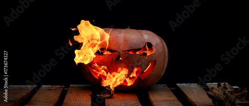 A very dangerous dangerous Halloween pumpkin, with a stern gaze and a smirk of a villain, in the darkness on a wooden pallet, spouts out the mouths and eyes of the flames of fire.