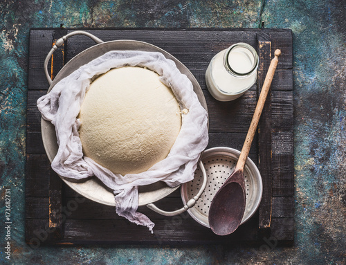 Homemade fresh cheese in  dish and cheesecloth with milk and wooden spoon on rustic background, top view photo