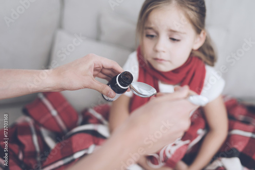Little sick girl sits on a white couch wrapped in a red scarf. She pours the syrup into the spoon