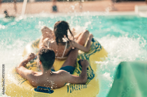 Fototapeta Naklejka Na Ścianę i Meble -  Cool people having fun on the water slide with friends and familiy in the aqua fun park glides playing happy and water splashes are all over. Blue sky background looks amazing sunlight