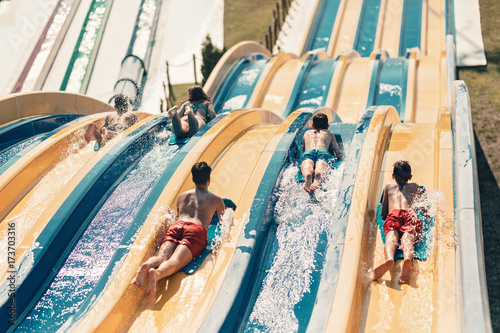 Young people having fun on the water slide with friends and familiy in the aqua fun park glides, happy falling into water and water splashes are all over. Blue sky background looks amazing sunlight photo