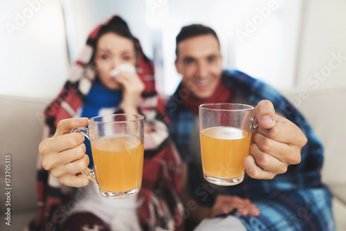 The couple is sitting on the couch wrapped in blankets. Man and woman are sick. They keep a cup of medical tea