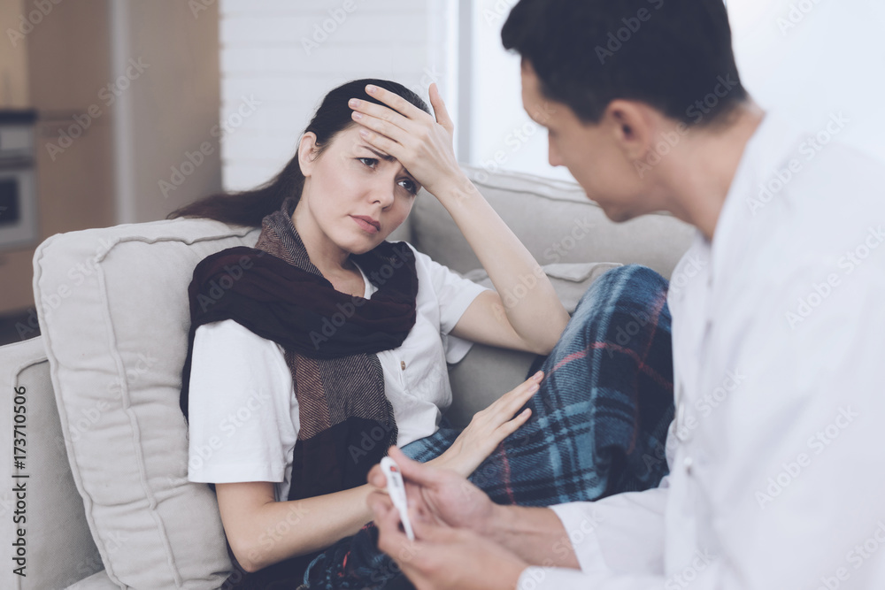 The woman fell ill and called the doctor at home. She sits on the sofa with a blanket. The doctor holds a thermometer The woman fell ill and called the doctor at home. She sits on the sofa with a blanket. The doctor holds a thermometer