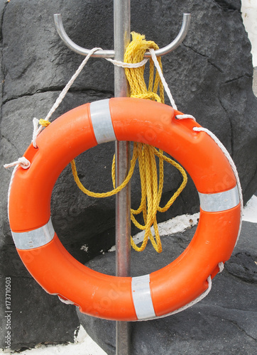 orange lifebelt or life preserver with yellow rope attatched on a stand in a resort swimming pool or harbour against grey rocks