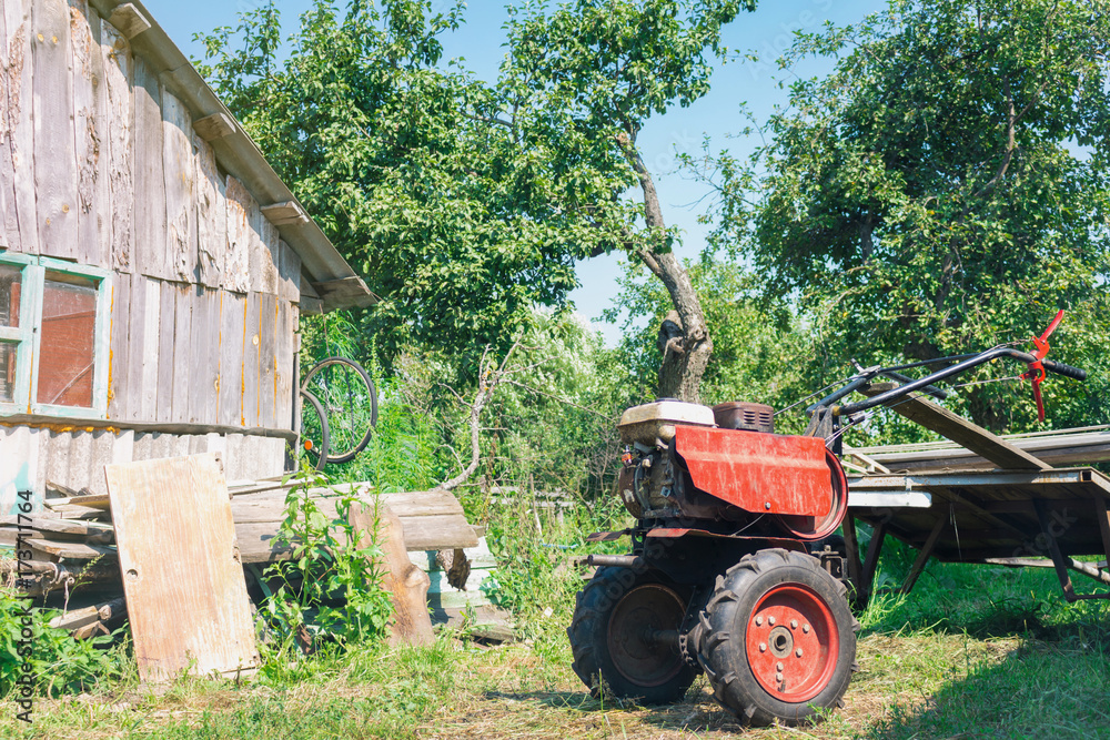Lawn mower in a village with green trees, a cart with firewood