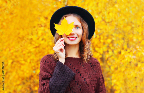 Autumn portrait smiling woman hides her eye maple yellow leaves in black round hat
