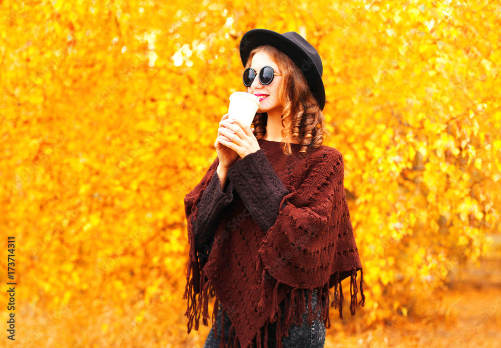 Fashion autumn woman drinks coffee in black round hat, knitted poncho over yellow leaves background Fashion autumn woman drinks coffee in black round hat, knitted poncho over yellow leaves background
