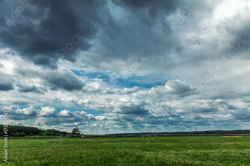 Fluffy rain clouds flying on a bright blue sky over a green field. Countryside landscape © Victoria Kondysenko