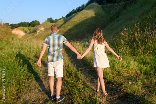 Young loving couple walking on green field near hills, free space. Young beautiful couple of travelers running near hill, back view. Stylish man and woman rest in country side outdoors