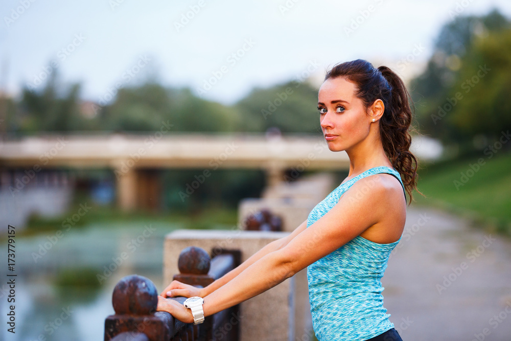 Portrait of young sporty woman before jogging in the evening city. Portrait of athletic girl in blue top relaxing after running at sunset