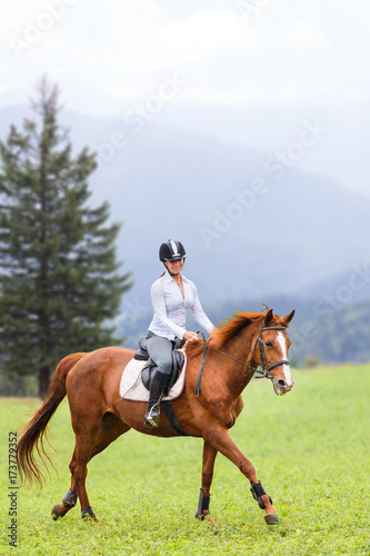 Young woman riding sorrel horse on green mountain meadow. Equestrian activity background