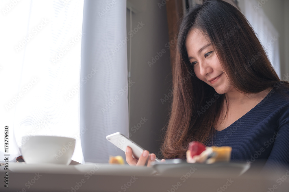Closeup image of a smiley beautiful Asian woman holding , using and looking at smart phone with white coffee cup and dessert plate on wooden table in vintage cafe Closeup image of a smiley beautiful Asian woman holding , using and looking at smart phone with white coffee cup and dessert plate on wooden table in vintage cafe