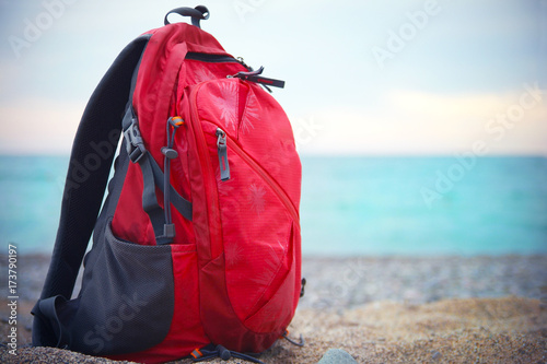In focus red backpack for traveling stands on a sandy sea shore on the background of blurred sea. Close up view.