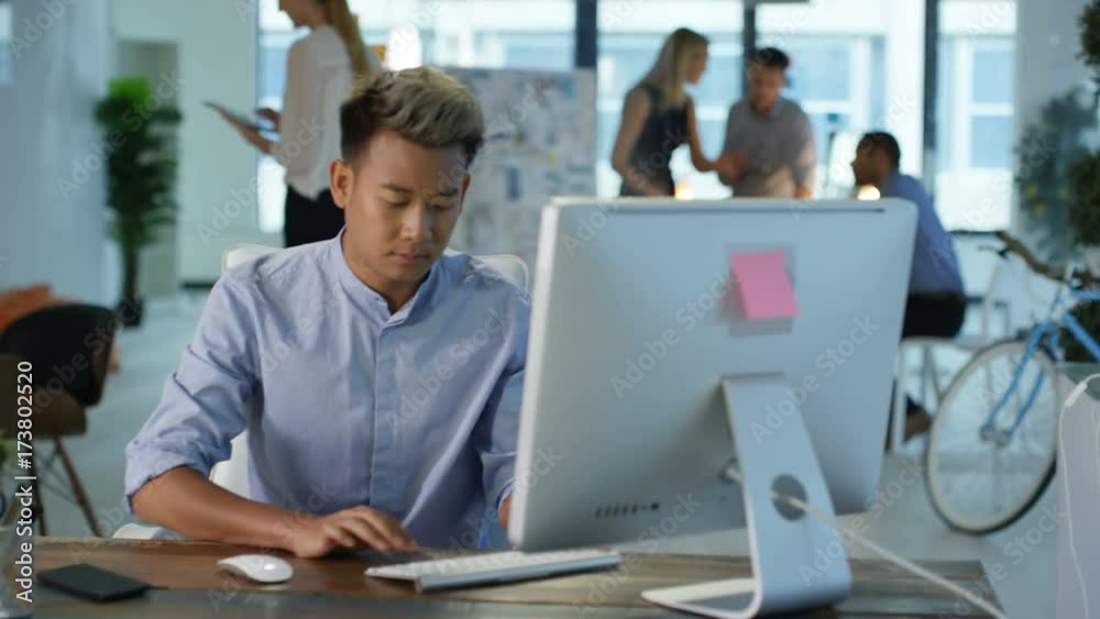 Portrait smiling designer at his desk with colleagues in a meeting in background. May depict third party? Portrait smiling designer at his desk with colleagues in a meeting in background. May depict third party?