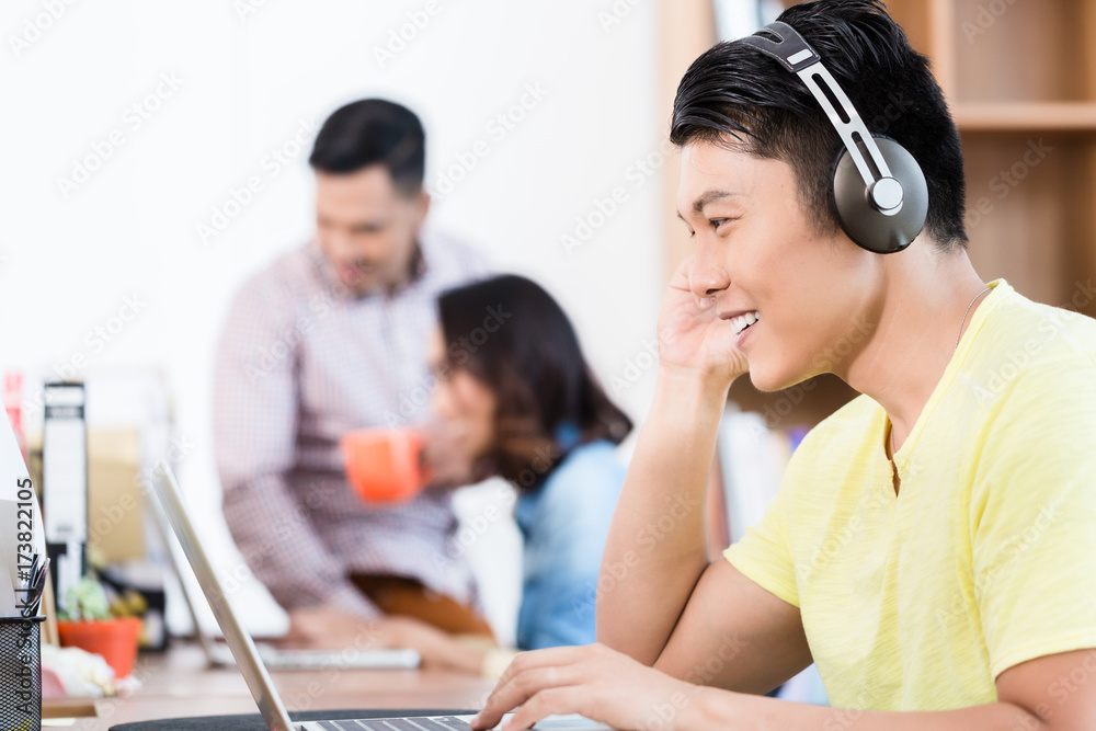 Side view portrait of a happy young Asian employee using headphones while watching a video presentation on laptop in the office