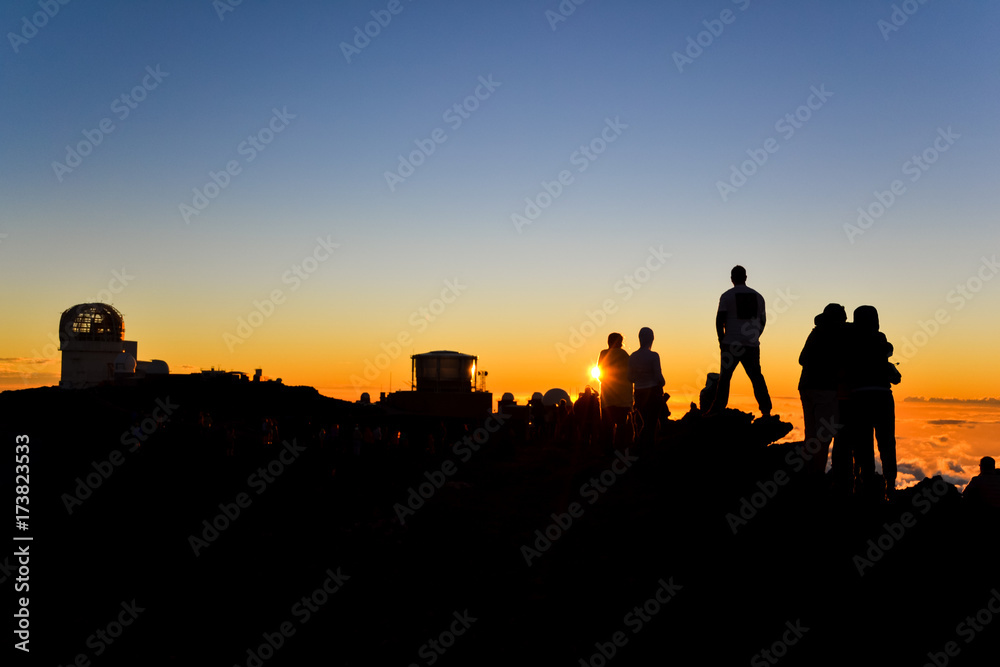 Tourists watching the spectacular sunset on the summit of Haleakala Crater (10,023 ft) on the island of Maui, Hawaii. Sun setting next to a spectator. Observatories in the background. 