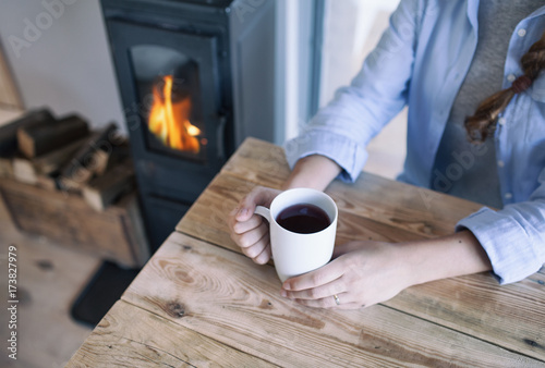 Woman with cup of coffee sitting by the fireplace and wooden table