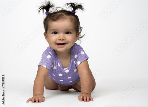 Smiling, infant girl crawling on white background dressed in purple, polka dot onesie