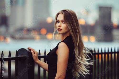 Fototapeta Gorgeous young model woman with perfect blonde hair looking at camera posing in the city wearing black evening dress