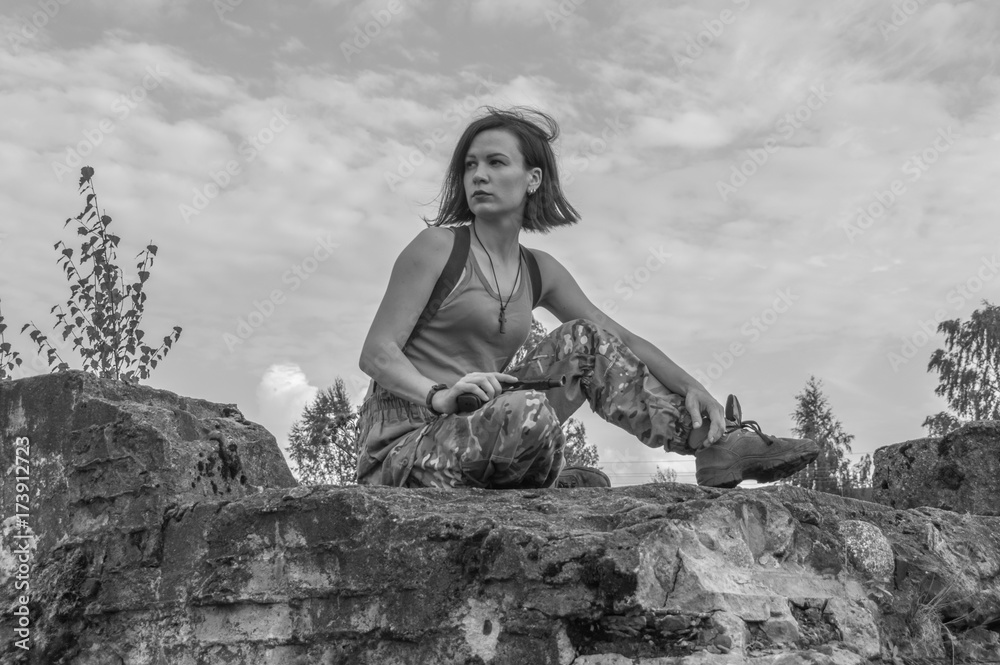 black and white, girl with a pistol in hand sitting on a large stone.