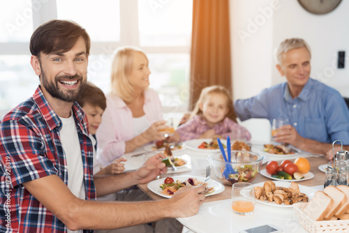 A man is posing and looking at the camera on the background of a family that sits at a festive table for Thanksgiving