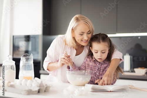 A girl with her grandmother is making a cake. A woman hugs a girl, together they read a book of recipes
