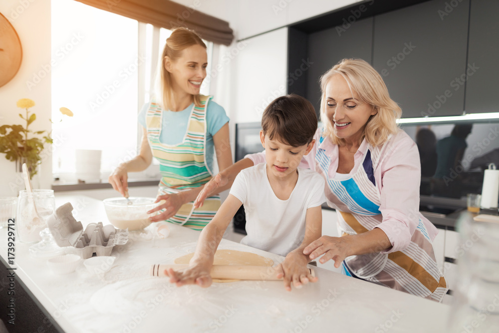 A woman teaches her son to cook homemade cakes. Her mother helps her. The boy rolls the dough with a rolling pin A woman teaches her son to cook homemade cakes. Her mother helps her. The boy rolls the dough with a rolling pin