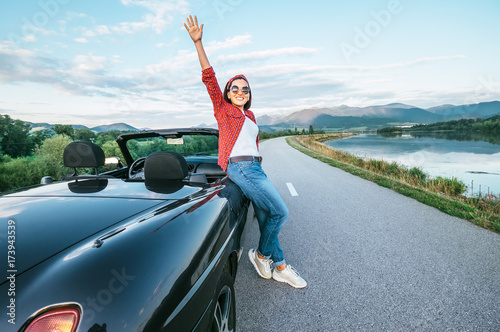 Happy smiling young woman stay near the cabriolet car on the mountain road