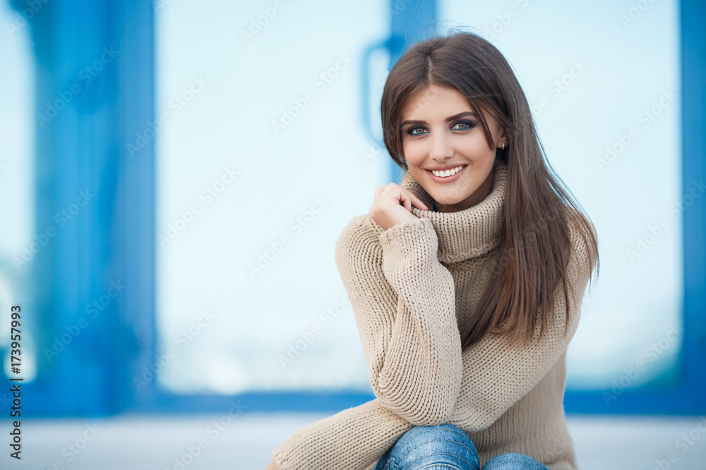 Obraz premium Young beautiful woman brunette with long straight hair and gray-green eyes,dressed in a beige sweater and blue jeans,hanging out in the city in the spring,sitting on the steps,one in the fresh air
