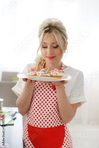 beautiful woman,with long blonde curly hair,dressed in a white t-shirt with short sleeves and white apron for the kitchen with red polka dots,cooks in the kitchen cupcakes with strawberries