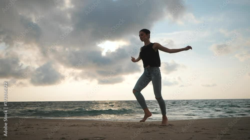 Young woman in casual style - jeans and black top doing ballet at the beach. Attractive ballerina practices in jumping on sandy coastline in autumn. Slow motion. Young woman in casual style - jeans and black top doing ballet at the beach. Attractive ballerina practices in jumping on sandy coastline in autumn. Slow motion.