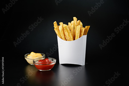 French fries in a white paper box with ketchup and cheese sauce isolated on black background. Front view.