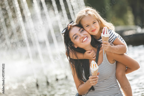 Young pretty mother and her daughter having fun together near the fountain. Beautiful woman and her little child eating ice cream. Cheerful family having fun.