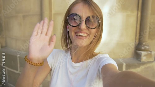Attractive caucasian fair hair girl in stylish sunglasses is having a video chat in front of an old building in the background, sunny day, slow motion