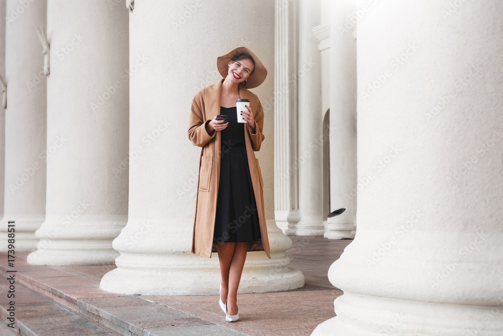 Girl in a brown coat a brown hat is walking and posing in the city interiors. The girl is smiling, checking her smartphone and drinking coffee from take away cafe. The photo looks very positive cause