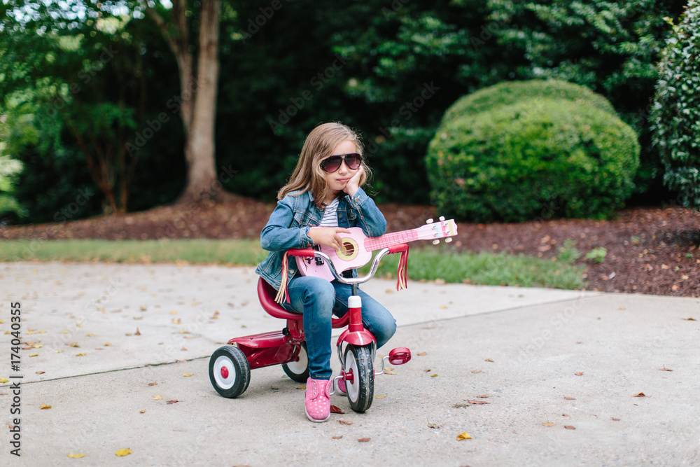 Cute young girl sitting on a tricycle while rocking out with her toy guitar Cute young girl sitting on a tricycle while rocking out with her toy guitar