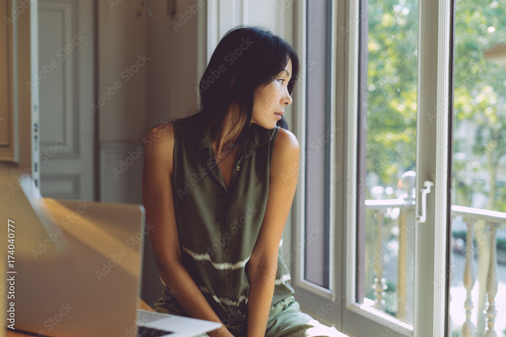 Attractive asian woman is looking through the window during break at the office. Business woman is thinking on the new project and making notes on a laptop. Student girl is sitting with a computer. Attractive asian woman is looking through the window during break at the office. Business woman is thinking on the new project and making notes on a laptop. Student girl is sitting with a computer.