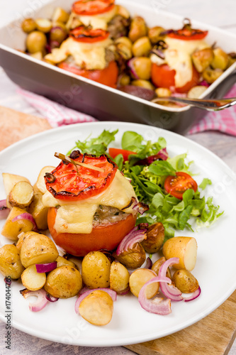 Oven baked tomatoes stuffed with minced meat, cheese and herbs. Served with new potatoes and salad. Casserole oven dish visible on white wooden background
